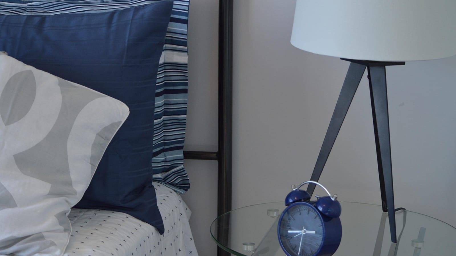 Bedside table with a lamp and a blue alarm clock next to a pillow