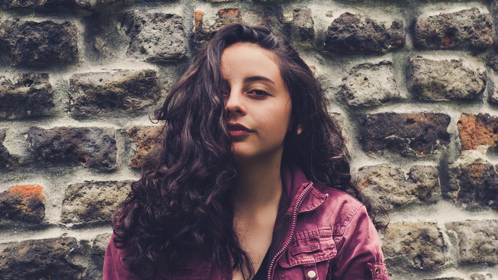 Young woman with long dark curly hair standing against a brick wall