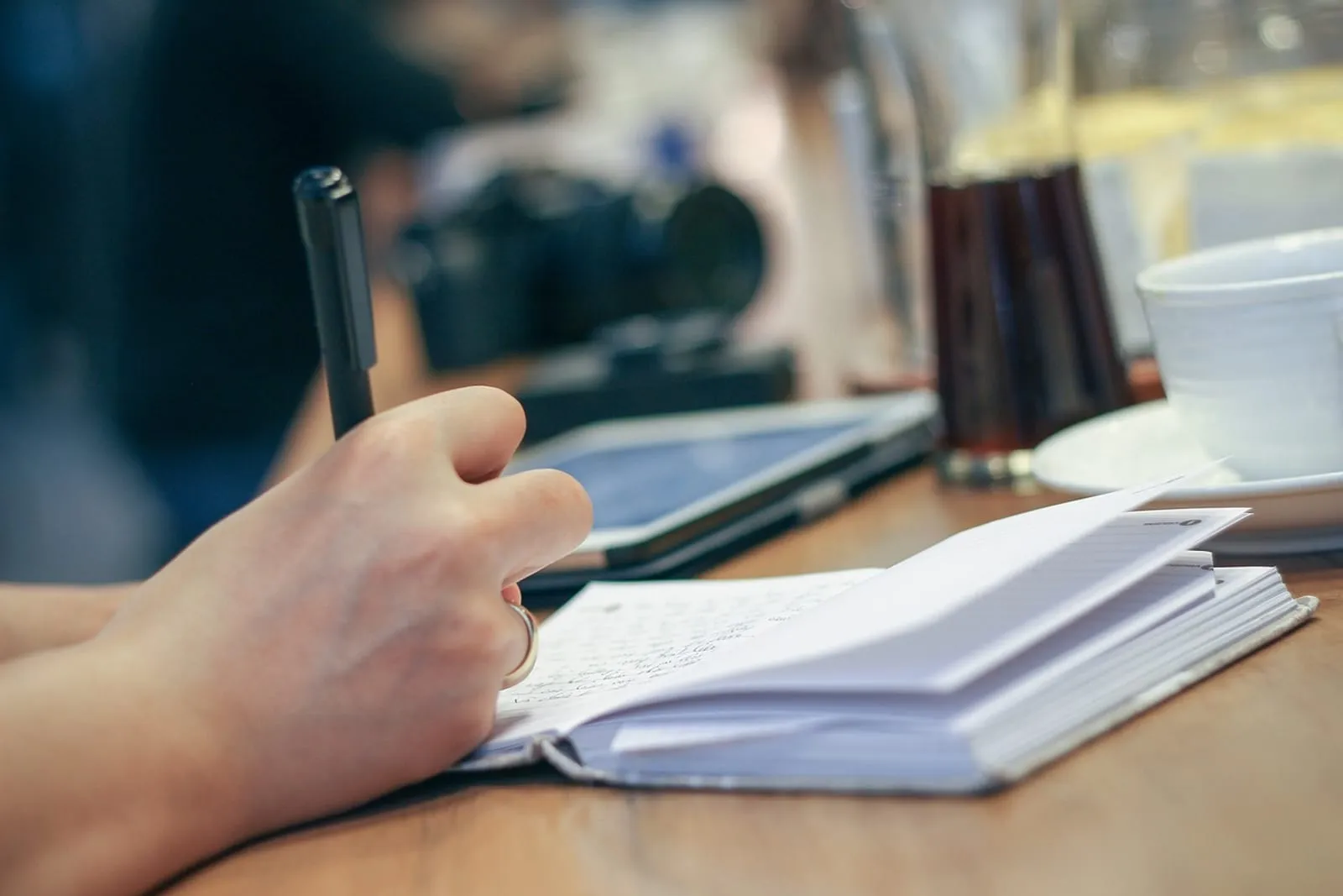 Hand writing in a notebook at a desk beside a tablet