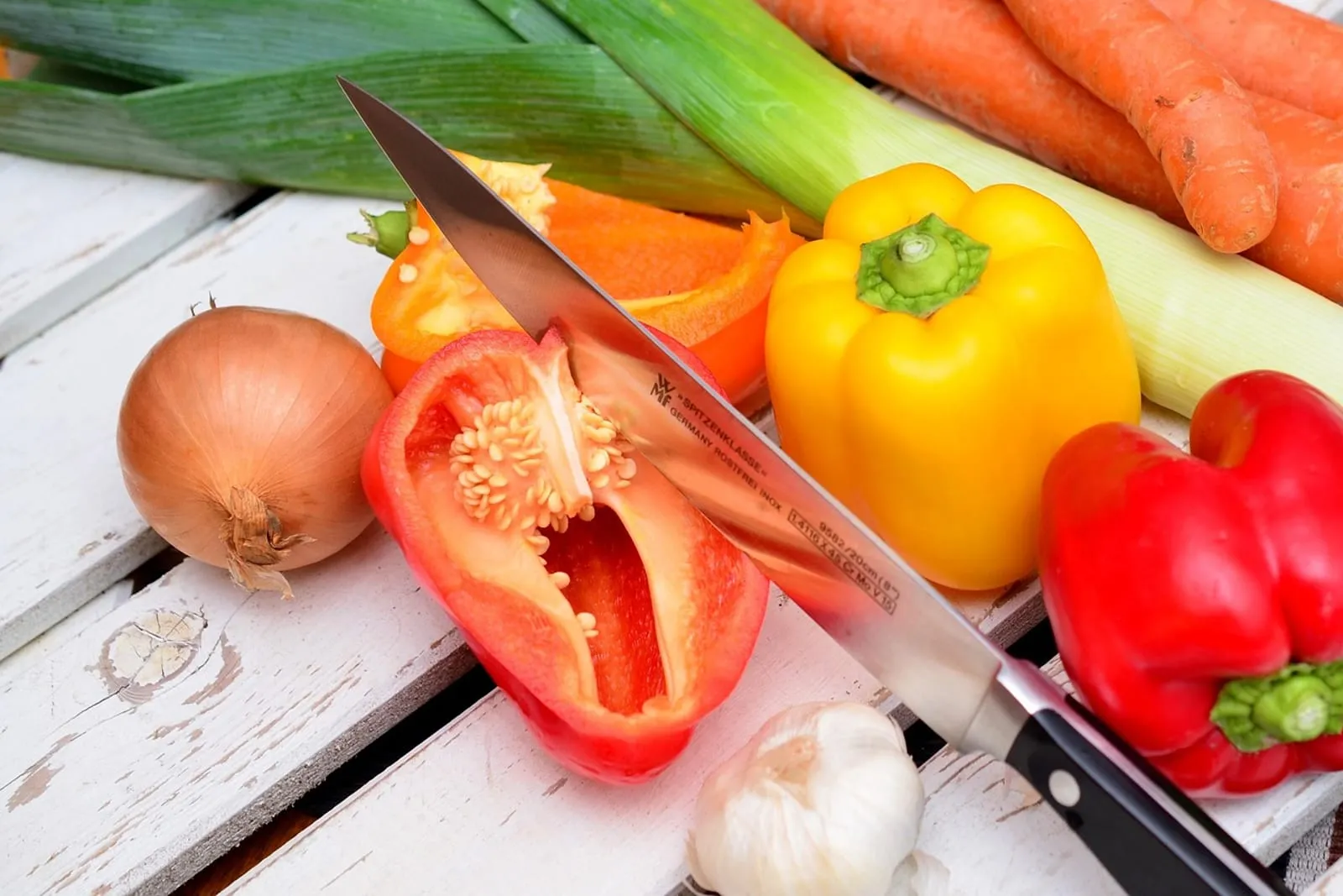 Chef's knife slicing a red bell pepper beside leeks, carrots, an onion and garlic