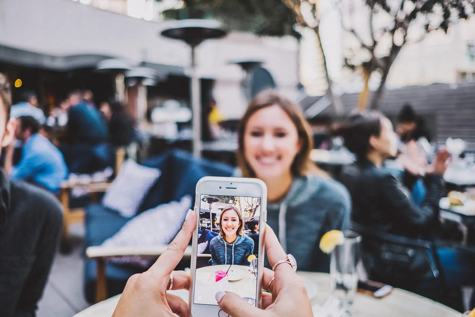 Person taking a photo of a smiling woman with a smartphone at an outdoor cafe